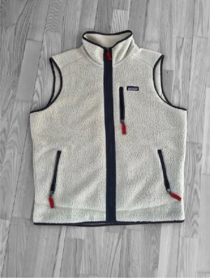 Beige fleeceväst från Patagonia - Snygg beige fleeceväst från Patagonia med svarta detaljer och röda dragkedjor. Västen har hög krage, tre fickor med dragkedja och är tillverkad i mjukt teddyfoder. Perfekt för lager-på-lager och outdoor-stil.