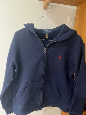 Mörkblå hoodie från Polo Ralph Lauren - Mörkblå hoodie med dragkedja från Polo Ralph Lauren. Klassisk design med huva, kängurufickor och den ikoniska röda logotypen broderad på bröstet. Tillverkad i mjukt bomullsmaterial som är skönt att bära. Perfekt för en avslappnad och stilren look. L i barn storlek (14-16)