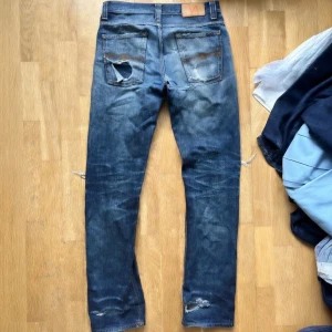 Nudie Jeans Grim Tim  - Säljer ett par blå jeans med snygga slitningar och fades. Jeansen har klassisk femficksdesign och raka ben. Perfekt för dig som gillar en avslappnad look och ett par jeans som är väldigt trendiga just nu. Jeansen är lagade i skrevet och kan behöva förstärkning snart igen. Storlek W31 L34. Hör av dig vid minsta fundering!