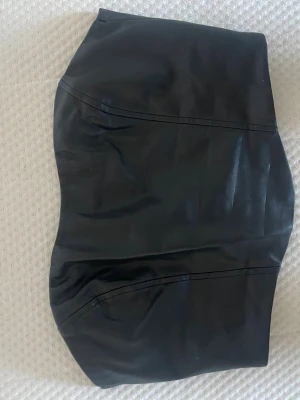 Svart tubtopp i skinnimitation - Snygg svart tubtopp i skinnimitation med dragkedja bak. Toppen har en bandeauformad passform och är perfekt för att skapa en edgy look. Materialet är mjukt och följsamt syntetmaterial som ger en cool känsla.