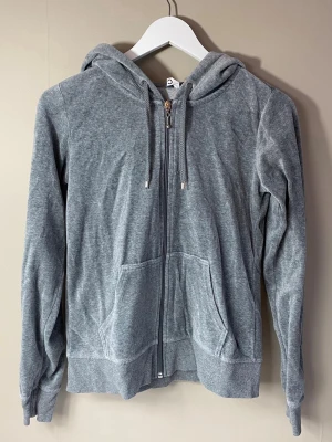 Grå zip hoodie med fickor - En stilren grå hoodie med dragkedja framtill och två fickor. Tröjan har huva med dragsko och ribbade muddar vid ärmslut och nederkant. Perfekt för en avslappnad look och enkel att matcha med andra plagg.