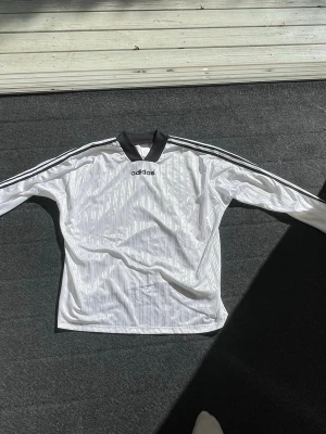 Vit långärmad tröja från Adidas - Säljer en vit långärmad sporttröja från Adidas med svarta detaljer. Tröjan har klassiska svarta ränder längs ärmarna, svart krage och muddar samt Adidas-logga på bröstet. Tillverkad i ett lätt och ventilerande material, perfekt för träning eller sport.