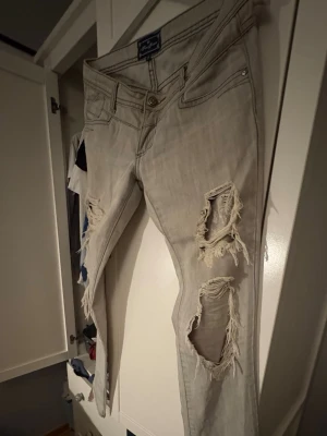 Ljusgrå slitna jeans  - Säljer ett par ljusgrå jeans från Replay med flera stora slitningar och hål på både fram- och baksida av benen. Jeansen har klassisk femficksmodell, låg midja och raka ben. Materialet är mjukt denim som ger en avslappnad look.