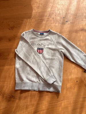 Grå sweatshirt från GANT - Snygg grå sweatshirt från GANT med broderad logga och amerikanskinspirerat sköldmärke på bröstet. Tröjan har rund hals, ribbade muddar och är tillverkad i mjukt sweatshirtmaterial. Perfekt för en avslappnad och stilren look. Den är aldrig använd 