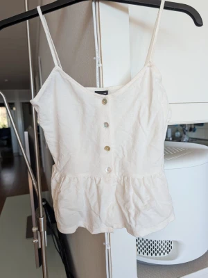 Cremevit linnetopp från Gina Tricot - Säljer en söt vit linnetopp från Gina Tricot i storlek XXS. Toppen har tunna axelband, dekorativa knappar framtill och en lätt rynkad nederkant som ger en avslappnad look. Perfekt för varma dagar och enkel att styla med jeans eller kjol.