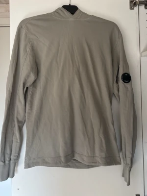 Beige hoodie från C.P. Company - Säljer en stilren beige hoodie från C.P. Company med klassisk huva och lång ärm. På vänster ärm sitter det ikoniska svarta locket med logga. Hoodien är tillverkad i mjukt bomullsmaterial och har ribbade muddar vid ärmsluten. Perfekt för en avslappnad och trendig look. Det finns ett lite hål som syns på bild 3 men inget som syns tidligt.