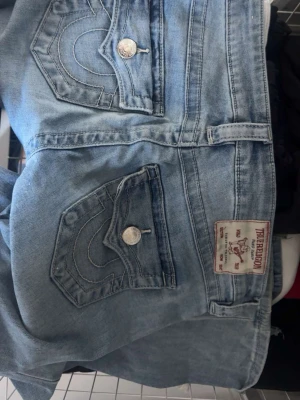 Ljusblå True Religion jeans - Säljer ett par ljusblå jeans från True Religion med klassiska kontrastsömmar och snygga fickdetaljer med silverfärgade knappar. Det hade ett litet hål i ena benet men sytt igen så syns knappt. Perfekta för en avslappnad och trendig look.