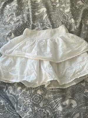 Vit broderad skort med volanger - Supersöt vit skort med broderade blommor och dubbla volanger. Resår i midjan för skön passform och luftig känsla. Perfekt för en somrig och avslappnad look. Materialet känns lätt och andas bra.