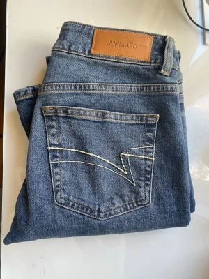 Blå bootcut jeans från Junkyard - Säljer ett par klassiska blå bootcut jeans från Junkyard. Jeansen har en snygg mellanblå tvätt, fem fickor och dekorativ söm på bakfickan. Tillverkade i slitstark denim med skinnpatch i midjan. Perfekta för en avslappnad och trendig look. Nypris 600kr