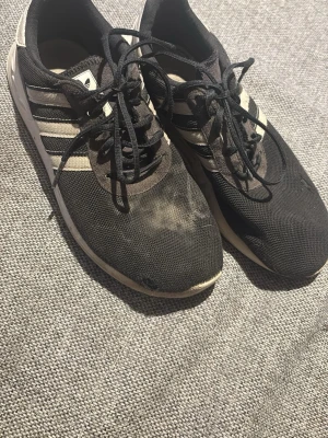 Svarta Adidas sneakers med vita ränder - Säljer ett par klassiska svarta Adidas sneakers med vita ränder på sidorna och vit sula. Skorna har mesh-överdel och snörning framtill. Perfekta för dig som gillar sportig och enkel stil.