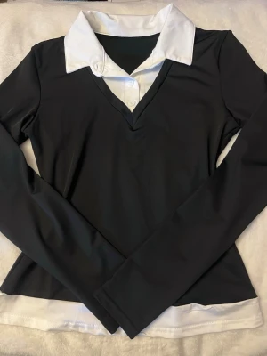 Svart långärmad topp med vit krage - Stilren svart långärmad topp med vit skjortkrage och vita knappdetaljer framtill.