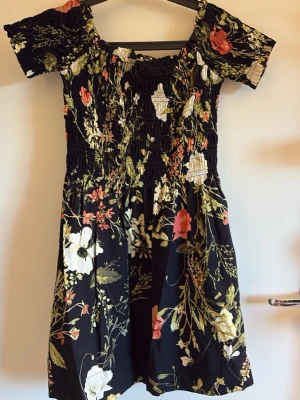 Svart blommig kortklänning - Supersöt svart kortklänning med blommigt mönster i vitt, grönt och rött. Klänningen har smock över byst och midja, korta ärmar och offshoulder-modell. Perfekt för sommarens alla tillfällen och ger en romantisk vibe. Stretchig.