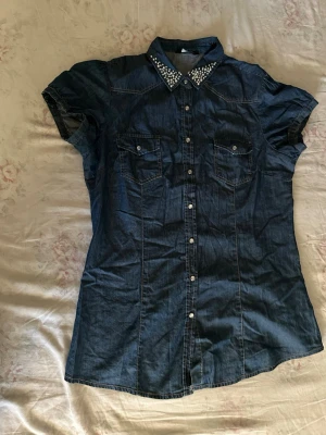 Mörkblå jeansskjorta med pärlor - Snygg mörkblå jeansskjorta med korta ärmar och pärlprydd krage. Skjortan har två bröstfickor med knappar och klassisk knappstängning framtill. Perfekt för dig som vill ha en cool och trendig look.