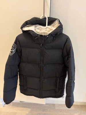 Svart dunjacka med huva och patch - Snygg svart dunjacka med stor huva och vit insida. Jackan har en patch på vänster ärm, två stora fickor framtill och dragkedja med vindslå. 