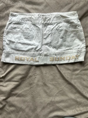 Vit vintage jeanskjol från 2000-talet - Vit kortkjol från Royal Sunday i bomull med broderad text längst ner. Kjolen har fickor fram, bälteshällor och stängs med dragkedja och knapp. Enkel och clean design som passar till många olika stilar.