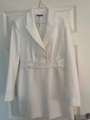 Vit klänning från Zara med bälte - Stilren vit klänning från Zara med klassisk krage och avtagbart bälte i midjan. Klänningen har en rak passform och långa ärmar, perfekt för en trendig och elegant look. Tillverkad i ett mjukt tyg som ger en lyxig känsla.