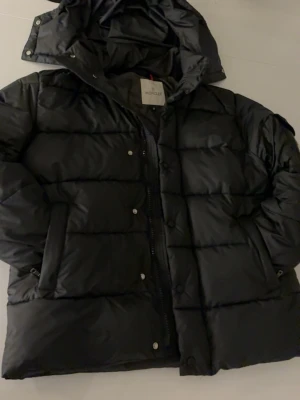 Svart dunjacka från Moncler - Snygg svart dunjacka från Moncler med huva och klassisk logotyp på ärmen. Jackan har quiltad design, dragkedja och tryckknappar framtill samt praktiska fickor. Perfekt för kalla vinterdagar och riktigt varm.