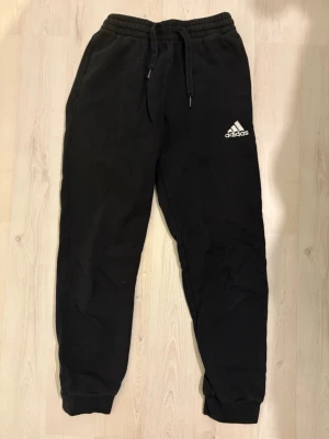 Svarta mjukisbyxor från Adidas - Svarta mjukisbyxor från Adidas med klassisk vit logga på vänster lår. Byxorna har resår i midjan med dragsko och muddar vid bensluten. Tillverkade i mjukt och bekvämt material, perfekta för chill eller träning.