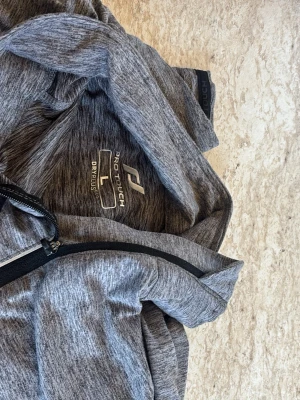 Grå träningsjacka Pro Touch - Sportig grå träningsjacka från Pro Touch med halv dragkedja framtill och hög krage. Tillverkad i funktionsmaterialet DRY PLUS som transporterar bort fukt. Perfekt för löpning eller gym. Snygg melerad design och mjuk känsla.