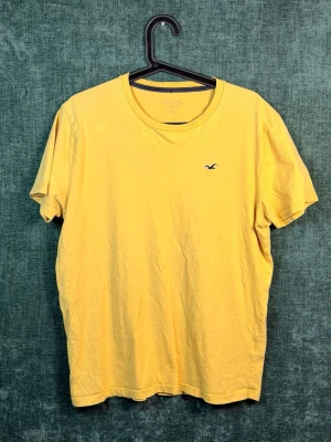 Gul t-shirt från Hollister - Snygg gul t-shirt från Hollister med klassisk rund hals och korta ärmar. Liten broderad logga på bröstet. Tillverkad i mjuk bomull som känns skön mot huden. Perfekt för en avslappnad och stilren look.