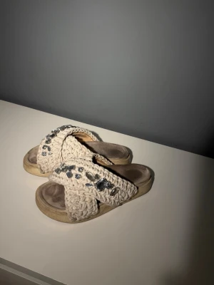 Beige sandaler med stickade band - Säljer ett par beige sandaler med breda, stickade korsade band och dekorativa stenar på ovansidan. Sandalerna har en platåsula i ljus färg och en mjuk innersula för extra komfort. Perfekta för en avslappnad och trendig sommarlook.