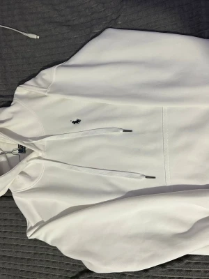 Vit hoodie från Polo Ralph Lauren - Säljer en stilren vit hoodie från Polo Ralph Lauren med klassisk huva och dragsnören. Tröjan har en broderad logga på bröstet och en stor magficka. Tillverkad i mjukt bomullsmaterial som är skönt mot huden.