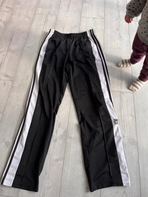 Svarta Adidas snap pants med ränder - Säljer ett par svarta Adidas mjukisbyxor med vita sidor och tryckknappar längs benen. De har använts fåtal gånger. Storlek S/M. :).    +Frakt 61kr