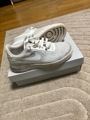 Vita Nike Air Force 1 Flyknit sneakers - Säljer ett par vita Nike Air Force 1 Flyknit sneakers med klassisk låg siluett. Skorna har stickad ovandel, vita skosnören och den ikoniska swoosh-loggan på sidan. Sulan är vit med AIR-detalj på hälen. Perfekta för dig som gillar stilrena och bekväma sneakers.