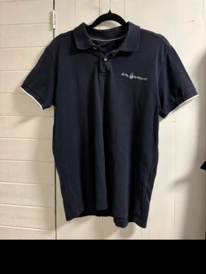 Marinblå pikétröja från Sail Racing - Klassisk marinblå pikétröja från Sail Racing med vit logga broderad på bröstet. Tröjan har korta ärmar, krage med knappar och vita detaljer på krage och ärmslut. Perfekt till en avslappnad och sportig stil.