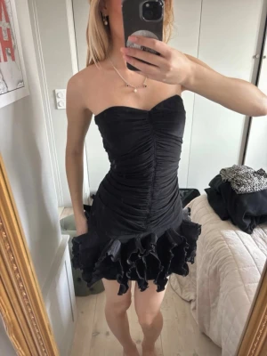 Svart axelbandslös volangklänning - Supersnygg svart kort klänning med axelbandslös design och volangkant nedtill. Klänningen har en åtsittande passform med rynkade detaljer framtill som ger en riktigt trendig look. 