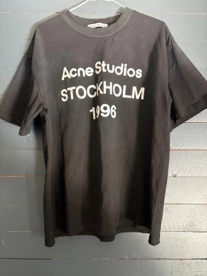 Svart Acne Studios t-shirt - Cool svart t-shirt från Acne Studios med vit text 'Acne Studios STOCKHOLM 1996' på bröstet. Klassisk rund halsringning och korta ärmar. Tillverkad i mjuk bomull för en skön och avslappnad look. Mindre L.