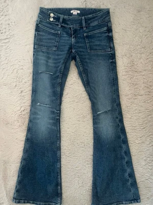 Låga Blå bootcut jeans med slitningar - Säljer ett par blå bootcut jeans i nyskick, aldrig använda. Jeansen har två framfickor, dubbla knappar i midjan och en klassisk tvättad look. Perfekta för en avslappnad och trendig stil. 