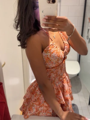 Blommig kort klänning med volanger - Supersöt kort klänning med blommigt mönster i orange, vit och rosa. Klänningen har smock i midjan, volangkjol och tunna axelband. Perfekt för varma dagar och ger en riktigt somrig vibe. Materialet känns lätt och luftigt.🌸☺️