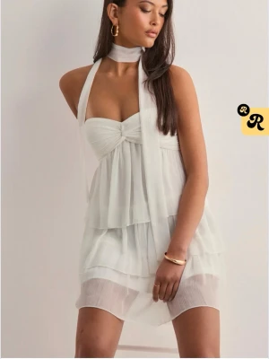 Vit omlott kortklänning med volanger - Supersöt vit kortklänning med omlottdesign och flera lager volanger. Klänningen har halterneck med knyt runt halsen och en lätt, luftig känsla. Endast använd en gång och i jätte bra skick!! 