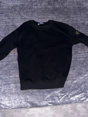 Svart sweatshirt från Stone Island - Säljer en svart sweatshirt från Stone Island med den klassiska loggan på vänster ärm. Tröjan har rund halsringning, ribbade muddar och är tillverkad i mjukt bomullsmaterial. Perfekt för en avslappnad och stilren look.