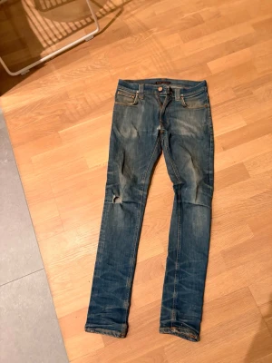 Blå slim fit jeans från Nudie Jeans - Säljer ett par blå slim fit jeans från Nudie Jeans med slitningar och hål på ena knät. Klassisk femficksmodell med dragkedja och knapp. Jeansen har en snygg tvättad look och kontrastsömmar. Perfekta för en avslappnad stil. 2 små hål. 