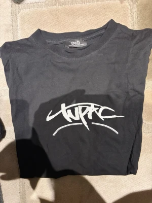 Svart Tupac t-shirt från D&G - Säljer en svart t-shirt från Dolce & Gabbana med stort Tupac-tryck framtill. T-shirten har en cool grafisk design med vit logga och färgglad illustration på framsidan. Klassisk rund hals och korta ärmar. Perfekt för dig som gillar streetwear och musikikoner.