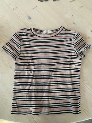 Randig ribbad t-shirt från H&M - Säljer en ribbad t-shirt från H&M med svart, vit och ljusrosa randigt mönster. T-shirten har korta ärmar och en klassisk rund halsringning. Har ett litet hål i armhålan men annars fint skick. Köparen står för frakt.