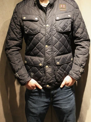 Vårjacka jacka från Morris - Snygg svart quiltad jacka från Morris med fyra fickor framtill, tryckknappar och dragkedja. Jackan har detaljer som spännen vid ärmsluten och en patch med brittisk flagga på bröstet. Perfekt för dig som gillar klassisk men modern stil. Modellen är 182cm