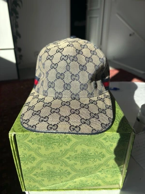 Gucci beige GG monogram keps - Säljer en beige keps från Gucci med klassiskt GG-monogram över hela kepsen. Kepsen har mörkblå insida och justerbart band bak. På sidorna finns Guccis ikoniska röd-blåa ränder. Materialet är canvas med detaljer i bomull. Perfekt för dig som vill ha en lyxig och trendig accessoar.