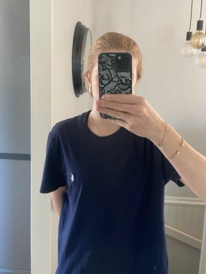 Marinblå t-shirt från Ralph Lauren - Klassisk marinblå t-shirt från Ralph Lauren med den ikoniska vita logon broderad på bröstet. T-shirten har rund halsringning och korta ärmar. Tillverkad i mjuk bomull för en skön och avslappnad look.