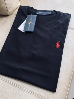 Polo Ralph Lauren T-shirt - Klassisk svart t-shirt från Polo Ralph Lauren med rund hals och den ikoniska röda logobrodyrn på bröstet. Tillverkad i mjuk bomull för en skön känsla och perfekt passform. Enkel och stilren design som funkar till allt.