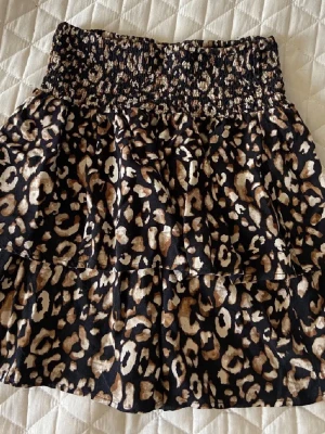 Leopardmönstrad kortkjol från Pigalle - Säljer en snygg kortkjol från Pigalle med leopardmönster i svart, beige och brunt. Kjolen har bred smock i midjan och volangdetaljer. Perfekt nu till sommaren. Osäker på storleken men passar mig som har storlek xs. Pris går att diskutera🥰