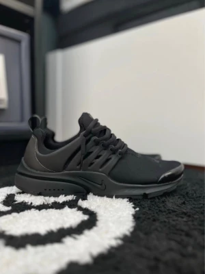 Svarta Nike Presto sneakers finns i 42-44 - Snygga helsvarta Nike Presto sneakers med låg profil och sportig design. Skon har en mesh- och syntetöverdel, rund tå och klassiska Nike-detaljer på sidan. Elastiska snören och bekväm sula för en modern look.
