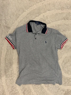 Grå pikétröja från Polo Ralph Lauren - Snygg grå pikétröja från Polo Ralph Lauren med marinblå krage och röda, vita detaljer på ärmsluten. Size M
