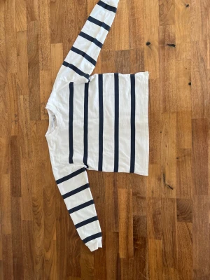 Vit långärmad randig tröja - Vit långärmad tröja med breda marinblå horisontella ränder över hela plagget, inklusive ärmarna. Klassisk rund halsringning och rak passform. Perfekt för en avslappnad och stilren look.