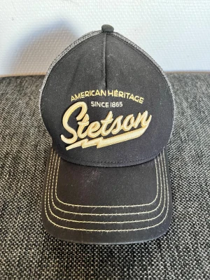 Svart Stetson keps med mesh - Säljer en svart keps från Stetson med gul brodyr framtill där det står 'American Heritage Since 1865 Stetson'. Kepsen har böjd skärm med kontrastsömmar och mesh på sidorna för extra ventilation. Perfekt för en avslappnad streetwear-look.
