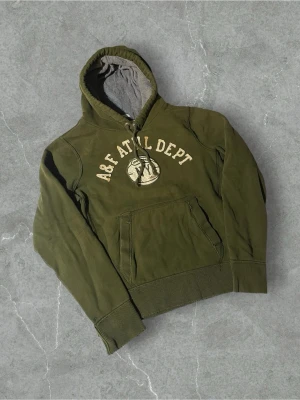 Olivgrön hoodie från Abercrombie | L - Olivgrön hoodie från Abercrombie & Fitch med broderad text 'A&F ATHL DEPT' och NY-logga på bröstet. Klassisk känguruficka, ribbade muddar och dragsko i huvan. Insidan av huvan är fodrad i grått tyg för extra komfort. Perfekt för en avslappnad streetwear-look.