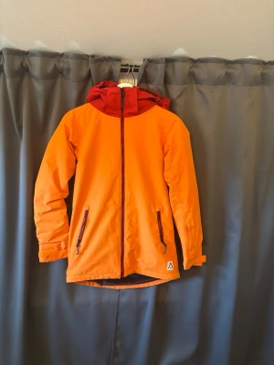 Orange skid/vindjacka med röd huva - Snygg orange skid/vindjacka med kontrasterande röd huva och dragkedja framtill. Två framfickor med dragkedja och justerbar huva. Jackan har en sportig look och är tillverkad i ett lätt, vindtätt material. Perfekt för blåsiga dagar och utomhusaktiviteter.