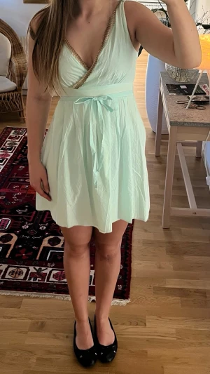 Kortklänning  - Söt turkos kort klänning med v-ringning och dekorativ kant i beige. Klänningen har omlott upptill, knytband i midjan och är ärmlös. Perfekt för varma dagar och har en luftig, lätt känsla.